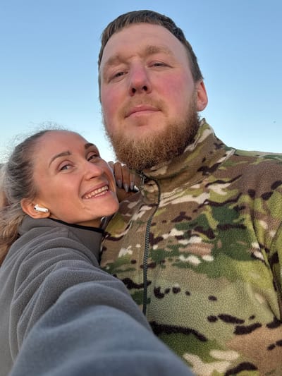 In Teriberka, Russia, a selfie shows a man and a woman in an outdoor setting. The man, with light brown hair and a beard, wears a camouflage fleece jacket. The woman, with blonde hair pulled back, is wearing a gray fleece jacket and a white wireless earbud. She is leaning her head on the man's shoulder and smiling at the camera. The background is a clear blue sky. The lighting suggests it is daytime.