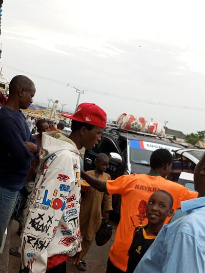 This image depicts a bustling scene, likely in the city of Gombe, Nigeria, possibly near a market or transportation hub. In the foreground, a young boy with a bright smile is the focal point, wearing a black and yellow sports shirt with the word "SPORTS" visible. He is standing next to an adult wearing a light blue shirt. To the left, a man in a dark blue long-sleeved shirt and jeans stands with his arms crossed, looking down. Another man, wearing a white hoodie adorned with colorful graffiti-style lettering, including words like "RASH" and "BATMAN," is the central figure in the middle ground. He wears a red baseball cap. Behind him, a dark SUV or van is visible, loaded with sacks on its roof, suggesting travel or transport. Several other individuals, including children and adults, are present in the background, some in traditional attire. The environment appears to be outdoors, with a cloudy sky overhead. The overall impression is one of everyday life and activity in an urban setting.