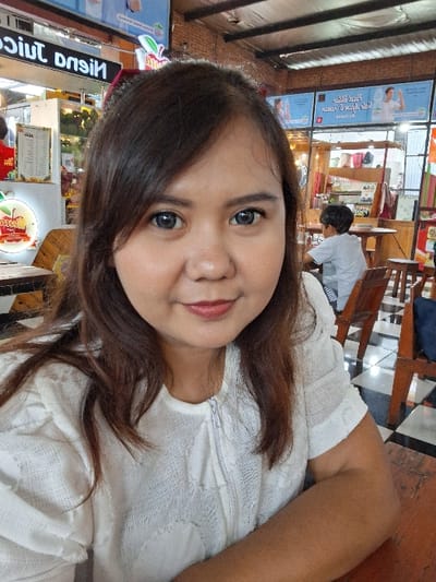 A woman with dark hair and fair skin is smiling at the camera. She's wearing a white textured top. The setting appears to be a casual restaurant or food court, as evidenced by the tables, chairs, and signage for "Nisnu Juice" in the background. A person in a gray shirt is visible in the distance behind her. The image is slightly rotated counterclockwise.