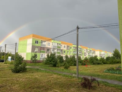 The image captures a suburban scene in Novinki, Russia, under a cloudy sky that has just produced a double rainbow. The foreground is dominated by a grassy area, with patches of dry, mowed grass and some areas of taller, green grass. A winding asphalt path cuts through the lawn, with puddles indicating recent rain.
In the mid-ground, a row of brightly colored apartment buildings stretches across the frame. The buildings are painted in alternating sections of lime green, white, and gray, with orange trim at the roofline. Many windows and balconies are visible, some with white railings. To the left of the buildings, several cars are parked, and a cylindrical kiosk or advertisement stands beside them.
A cluster of young trees is scattered throughout the grassy area in front of the buildings, with taller trees and shrubs forming a line behind them. A small, red metal swing set is visible on the grass to the right of the path. Further to the right, a small garden plot with raised beds and some flowering plants can be seen.
The sky is overcast, with the faint presence of a second rainbow above the more prominent one. The lighting suggests it is daytime, likely late afternoon or early evening after a rain shower. The overall mood is serene and peaceful, despite the overcast weather, with the vibrant colors of the buildings and the natural beauty of the rainbow adding a touch of optimism. The scene depicts a residential area, likely a newly developed one, with green spaces and amenities for its residents. No people are visible in the image, and no text is discernible.