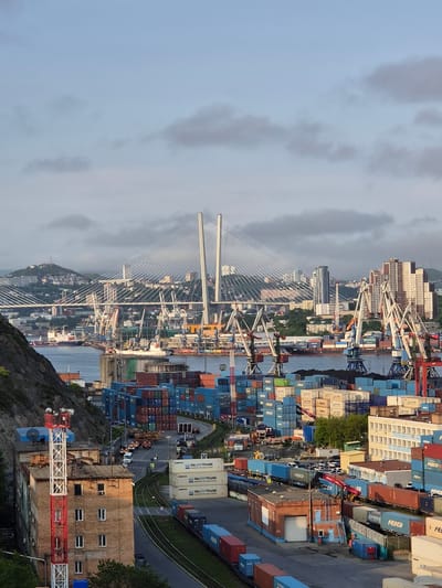 This is a high-angle, eye-level, long shot of a busy port in Vladivostok, Russia, dominated by a large cable-stayed bridge in the background. The port is filled with shipping containers stacked high and numerous cranes of various sizes, including large tower cranes and gantry cranes. Several cargo ships are visible in the water between the port and the bridge. In the foreground, there is a brick building with a red and white lattice tower structure attached to it, which appears to be an antenna or communication tower. Trains carrying containers are on tracks running through the port. The sky is overcast with scattered clouds. The overall scene depicts a bustling industrial maritime hub.