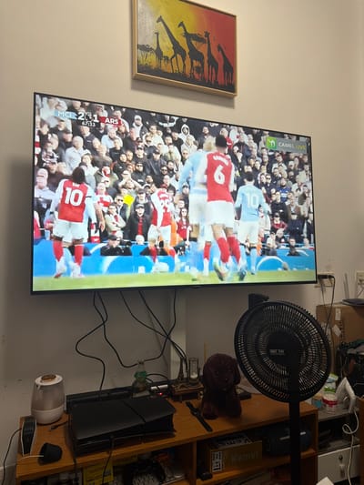 A television is on, displaying a soccer game between Manchester City (MCI) and Arsenal (ARS). The score is 2-1 in favor of Arsenal, with the game clock reading 67:33. Visible on the screen are players wearing the numbers 6 and 10. To the right of the TV is a black fan, shelves stacked with various items, including a Kemei box and a Sony PlayStation 5 console. Cords from the TV and other electronics are tangled. To the left of the TV is a framed painting or print featuring giraffes against a sunset. The word "SAST 先科" is visible on the fan. It is impossible to confirm the location in the city of Jiangning District, China from this image.