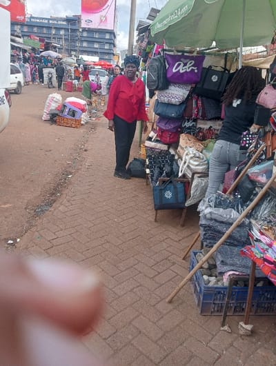 An outdoor market scene depicts a woman in a red blouse and black pants standing near a stall displaying handbags. Another person with long dreadlocks is seen browsing the bags. The stall is covered by a green umbrella. Behind them, a street scene with other vendors, pedestrians, and vehicles unfolds. In the foreground, a brick-paved ground and a blurry finger obstructing the view are visible. A building with a billboard is in the background.
