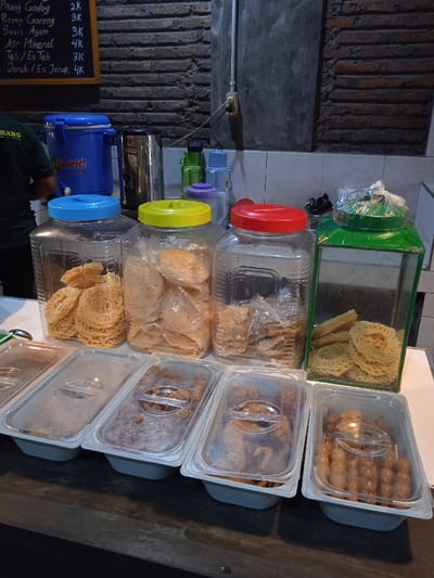 The image shows a food stall in Surakarta, Indonesia, located at coordinates -7.567 latitude and 110.799 longitude. The stall is situated in a brick-walled room. The focal point of the image is a row of five clear plastic containers filled with various foods. The first container holds a stack of fried pastry squares. The second container is filled with a similar type of pastry, but the squares are larger and more irregular in shape. The third container also holds a stack of fried pastry squares. The fourth container holds small, flat, round fried pastry. The fifth container holds what appears to be small, round, cooked snacks. The food items are arranged in a neat and organized manner. Behind the containers, there are three larger containers with lids, each filled with different types of fried snacks. These are arranged neatly on a counter. One of the containers has a blue lid with the word "Spirit!" printed on it. There is a partially visible figure on the left edge of the image. The figure is wearing a black shirt. The back wall of the stall is adorned with a blackboard displaying a price list in Indonesian. There is a wall-mounted switchboard on the wall. The scene is clean and well-lit. The lighting is artificial, coming from a source that is not visible in the image.