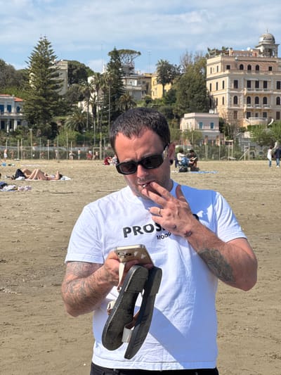 A medium shot captures a man holding a pair of sandals and smoking a cigarette on the beach in Anzio, Italy. The man, wearing sunglasses and a white t-shirt, is tattooed. The beach is sandy, and in the background, buildings and trees are visible. The scene is set during daylight hours. Other people are dispersed across the beach.