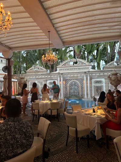 The image captures an outdoor dining scene at The Villa Casa Casuarina, located at 1116 Ocean Drive, Miami Beach, United States. The setting is an open-air covered patio beneath a striped fabric canopy from which two illuminated crystal chandeliers are suspended. A dark-colored ceiling fan is mounted on a structural beam to the left.
The floor is a dark-toned mosaic or cobblestone pattern. Several round dining tables, each covered with a white tablecloth, are arranged throughout the area. Each table is set with white plates, various glassware, and cutlery, with some featuring small condiment bottles. Guests are seated in white upholstered chairs with dark wooden frames.
In the background, an ornate, neoclassical-style wall made of light-colored stone and intricate mosaic tiling forms the perimeter of a blue-tiled swimming pool. A central fountain within the pool is active, emitting water jets, and features a mosaic design. Two large, decorative urns are positioned on top of the wall. Behind the wall, lush tropical vegetation, including large banana tree leaves, is prominent, with parts of a distant building visible through the foliage.
Approximately 15-20 individuals are present. In the mid-ground, a group of four stands near the pool's edge: a light-skinned male in a light blue collared shirt gestures with his right hand while facing three women. One woman wears a white strapless dress, one a dark sleeveless dress, and another a light-colored short-sleeved dress; each appears to be holding a mobile phone. Seated diners occupy the foreground and mid-ground. A male in the immediate foreground is visible from the back, wearing a dark patterned short-sleeve shirt. To the right, a woman with long dark hair, wearing a dark top, is observed looking at a menu, with another woman in a red top partially visible to her right, also examining a menu.