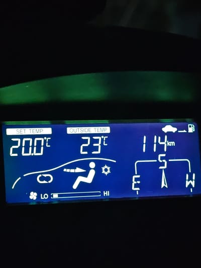 This is a close-up, full-frame, eye-level, indoor shot of a car's digital display. The display shows various information about the car's climate control and other functions. On the left, under "SET TEMP.", it reads "20.0°C". In the center, under "OUTSIDE TEMP.", it reads "23°C". To the right, it displays "114 km" and an icon of a car next to a gas pump. Below these readings are symbols related to air conditioning: a fan speed indicator showing "LO" and "HI", a circular arrow indicating air recirculation, an icon of a person with arrows indicating airflow direction, and a snowflake symbol. A compass rose is also visible with directional indicators E, S, and W. The background of the image is dark, with a faint green glow along the bottom edge, suggesting ambient light or a reflection within the car. No people are visible, and no specific location in Shumen, Bulgaria, can be identified from the image content.