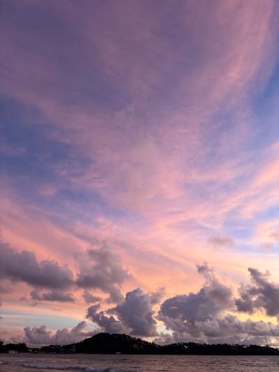 The image depicts a twilight sky over a body of water, likely a lake or ocean, with a silhouetted treeline and buildings along the distant shore. The sky is a dramatic canvas of colors, transitioning from a soft purple and pink on the left to warmer oranges and yellows on the right, where darker, textured clouds are clustered. Wispy, cirrus-like clouds are scattered across the purple expanse, catching the fading sunlight. The water in the foreground is rippled, reflecting the colors of the sky. The scene suggests the end of the day, with the warm hues indicating a sunset. The presence of buildings and trees suggests a populated area, possibly a coastal town or resort. The overall mood is serene and beautiful, evoking a sense of calm and natural wonder. There is no visible text in the image.