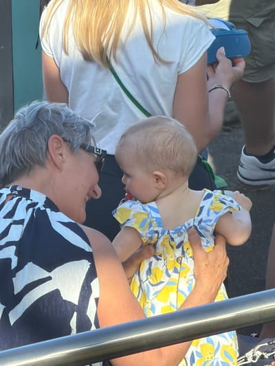 The image depicts three individuals in an outdoor setting. An infant in a yellow and blue floral dress is being held by an older adult wearing a black and white patterned shirt. The infant is facing away from the viewer. A third person, wearing a white shirt with a green strap and blue footwear, is partially visible to the left. The background includes other people and a railing. The location is not identifiable as Fentange, Luxembourg.