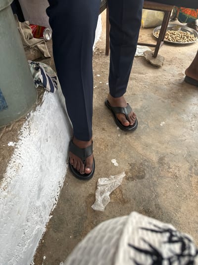 This is a close-up, eye-level, medium shot of a person's legs and feet, positioned in an outdoor setting in Narayi, Nigeria. The person is wearing dark navy trousers and black flip-flops. The ground is made of concrete with some scattered debris, including a crumpled piece of clear plastic. To the left, a white-painted wall is visible, showing signs of being freshly painted or plastered. Next to the wall, there's a light-colored bucket or container and a red Schweppes can with a silver opening. A piece of striped fabric lies on the ground near the bucket. In the background, on a metal stool, is a metal tray filled with shelled peanuts. Another person's feet, wearing sandals, are partially visible in the far right of the frame. The overall scene suggests a casual, everyday environment, possibly a market or a roadside stall.