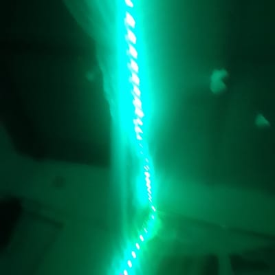 A blurry, angled view of bright green LED strip lights dominates the composition. The lights are a linear array of small, intense points, illuminating an otherwise dark space. Background elements, barely visible, suggest architectural features like walls and possibly a ceiling, all bathed in the green glow. The image lacks sharp focus, enhancing the atmospheric effect of the light.