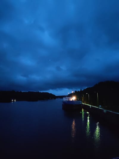 This is a night photograph of a body of water, possibly a canal or river, in the city of Devyatiny, Russia. The sky is dark blue with scattered clouds and a faint crescent moon. On the right side of the image, a treed embankment lines the water, with several streetlights illuminating the path. A barge is docked or transiting on the left side of the waterway, with some lights visible on its structure. The lights from the barge and streetlights are reflected on the dark water. In the distance, lights are visible along the shore on the left side of the image, suggesting the presence of buildings or homes.