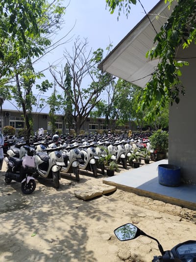 The image shows a motorcycle parking area at an unidentified location in Meunasahpantonlabu, Indonesia. Rows of white and light-colored motorcycles are parked in an orderly fashion. A few motorcycles of different colors are also visible. A building with a white roof and grey walls is present in the background and to the right of the frame. Trees surround the parking area, casting shadows on the ground. A blue plastic container sits on the ground near the building. A portion of another motorcycle is visible in the foreground. People are seen in the background near the building. The ground is dirt and shows evidence of foot and vehicle traffic. Plant pots are placed in-between some of the motorcycles.