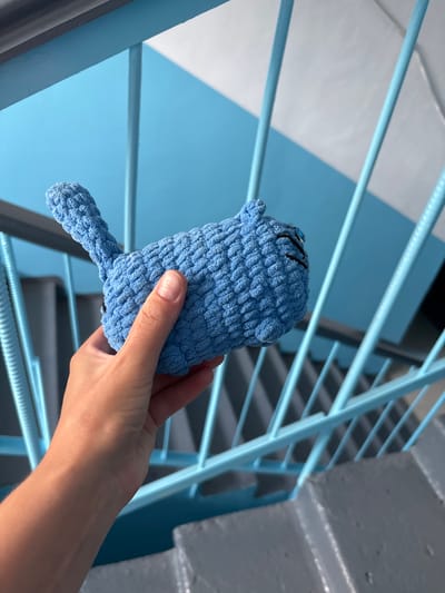 A person's hand is holding a small, light blue, crocheted toy animal, possibly a cat, in front of a set of light blue metal stairs. The toy has black embroidered eyes and eyelashes. The stairs are part of an outdoor staircase, with grey concrete steps visible below. The background consists of light blue and white panels, creating a diagonal pattern. The overall scene suggests an urban environment, potentially a public space in Blagoveshchensk, Russia.