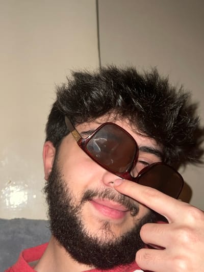 A person with dark hair and a full beard is wearing brown square sunglasses, held askew by a finger. The frames are a dark, reddish-brown color. The person has fair skin. The background is a plain wall, contrasting with the dark hair. A portion of what appears to be a red garment is visible. The lighting casts shadows and reflections on the sunglasses and face.