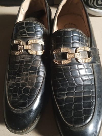 The image shows a close-up, overhead view of a pair of black, alligator-textured loafers with gold and diamond-encrusted embellishments. The shoes are dusty, indicating they may not be new or have been stored for some time. One shoe is slightly to the left and the other is slightly to the right, both positioned side-by-side on a neutral surface. The background is dark and indistinct, offering no specific environmental clues. There are no people visible in the image, nor any indication of the location being within Ashata, Nigeria.
