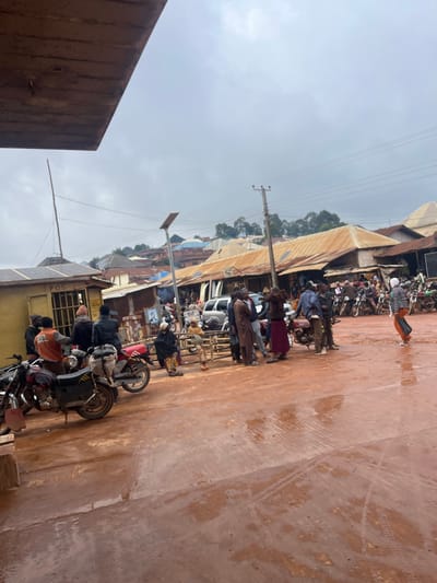 This is a sideways, slightly blurry, medium-shot photograph taken outdoors in what appears to be a marketplace or busy street in Gembu, Nigeria. The scene is dominated by a muddy, reddish-brown ground, suggesting recent rain or a dusty environment. A line of motorcycles, some with riders or passengers, are parked or moving along the street.
Several people are visible, some standing, some on motorcycles. They are dressed in casual clothing, including shirts, pants, and head coverings. Their expressions are not clearly discernible due to the image quality and distance, but the general atmosphere seems busy and perhaps a bit congested due to the parked vehicles and pedestrian activity.
In the background, there are various buildings with corrugated metal roofs, characteristic of many market structures in such regions. There are also utility poles and wires, indicating the presence of infrastructure. The sky is overcast, suggesting a cloudy or possibly rainy day.
One building has visible text that reads "POST" and another has a logo that appears to be "MTN". These offer clues about the types of businesses or services available in the area, such as postal services and a telecommunications provider.
The overall impression is of a bustling daily scene in a Nigerian town, with a focus on local transportation and commerce. The time of day is likely daytime, given the ambient light, but the overcast conditions make it difficult to pinpoint further.