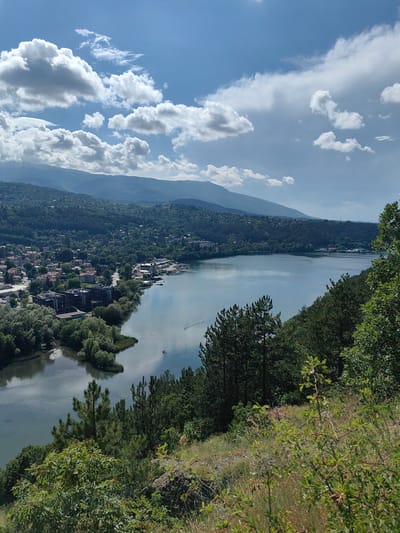 This image shows a wide-angle view of Pancharevo Lake in Pancharevo, Bulgaria. The lake is calm and reflects the sky and surrounding landscape. On the left side of the image, there is a town with buildings nestled amongst trees, leading up to a hill. The right side of the image is dominated by a dense forest that slopes down towards the lake. The sky is partly cloudy with dramatic white clouds against a blue sky. In the foreground, there is a grassy hillside with some bushes. The scene conveys a sense of tranquility and natural beauty.