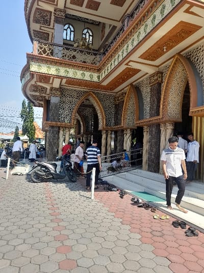 The image shows the exterior of a mosque in Kecamatan Candi, Indonesia, located at latitude: -7.494, longitude: 112.717. The mosque has an intricate facade with intricate details, including arches, columns, and decorative tile work. There are people standing in front of the mosque. Some are wearing traditional Muslim clothing, while others are wearing casual attire. A black motorcycle is parked in front of the mosque. The people are interacting with each other and appear to be waiting to enter the mosque. There are shoes and sandals lined up on the steps leading up to the mosque. The environment is sunny and bright.