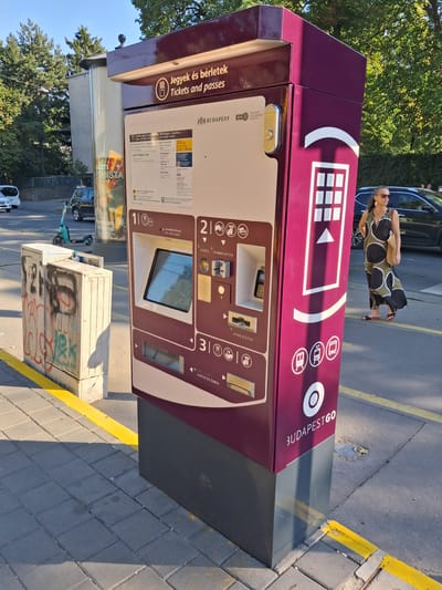 The image captures a Budapest public transport ticket vending machine on a sunny day. The machine, predominantly a deep maroon color with some beige panels, displays text in Hungarian, including "Jegyek és bérletek" (Tickets and passes). It features a touchscreen display, coin and banknote slots, and a ticket dispenser. Several icons representing different transport modes, such as a tram and a bus, are visible. A woman in a patterned dress is standing to the right of the machine, facing away from the camera, and appearing to be waiting. In the background, a street is visible with parked cars and trees. To the left of the ticket machine, there is a graffiti-covered utility box. The ground is paved with cobblestones and asphalt, with yellow lines indicating a pedestrian crossing or curb. The scene suggests a typical urban environment in Budapest.