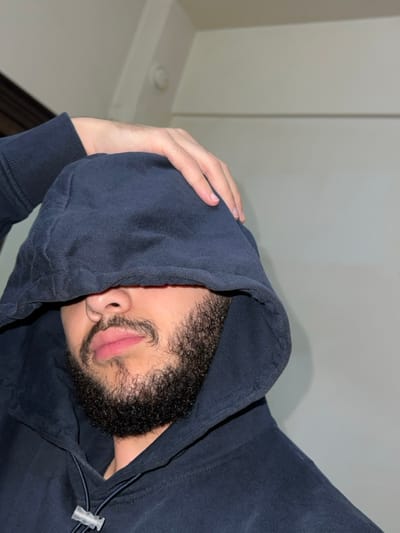 A person wearing a dark blue hooded sweatshirt is photographed from a low angle, obscuring their face. The hood is pulled over their head, covering most of their face, with only the mouth and beard visible. A hand is seen holding the hood near their head. The background features a plain white wall and ceiling corner. The photo has a casual and slightly mysterious vibe.