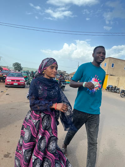 In Rafin Par, Nigeria, a man and a woman walk side by side on a dusty, paved road. The woman, wearing a black and pink patterned dress and headscarf, looks slightly away from the camera, her right arm adorned with a red bracelet. The man, in a teal t-shirt with a colorful paint-splatter design and dark jeans, faces the camera and smiles. He has a white wristband on his left arm. In the background, a red Volkswagen car, a green auto-rickshaw, and several motorcycles are visible, along with a two-story building with yellow and light-colored walls and blue-framed windows. The sky is a vibrant blue with scattered white clouds.