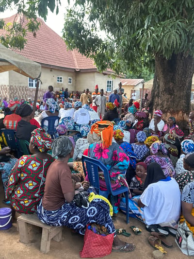 A crowd of people, predominantly women wearing brightly colored headscarves and wrappers, are gathered outdoors in a village setting. They are seated on chairs and the ground under the shade of a large tree, with a building with a red tiled roof in the background. The scene appears to be in Mararaba, Nigeria, likely for a community event or religious gathering. Some individuals are standing, including men in traditional attire, and one man in a purple robe appears to be speaking or leading. Objects in the scene include plastic chairs, wooden stools, a purple bucket, and various bags and footwear scattered on the ground. The atmosphere seems to be relaxed and communal.
