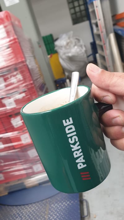 A hand holds a green ceramic mug with the white logo "PARKSIDE" and three red stripes, containing a coffee beverage with a spoon. The mug is in the foreground, with a blurred background of industrial shelving units, stacks of red boxes wrapped in plastic, and large white bags. The scene appears to be indoors, possibly a warehouse or storage area.