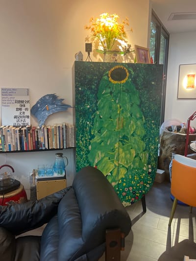 The image depicts an indoor scene, possibly a living room or study, with furniture and decorative items. The focal point is a tall, green cabinet featuring a large, stylized sunflower painting. To the left of the cabinet, a bookshelf is filled with books. Above the bookshelf, a blue, irregularly shaped clock is mounted on the wall. A framed picture and a bouquet of orange flowers with a spotlight are visible on top of the cabinet. In the foreground, a black leather sofa is partially visible. In the background, an orange chair and a large, textured urn-like object are present. The lighting suggests a warm, ambient environment. The presence of Chinese characters on some of the books and posters indicates a likely location in China, potentially within Liandu District. There are no people visible in the image.