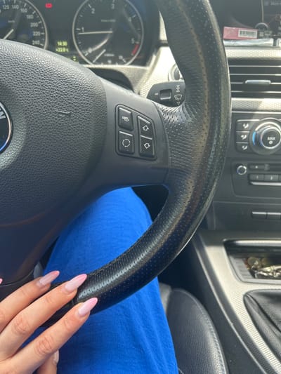 The image provides a close-up, first-person view from the driver's seat of a BMW car in Razlog, Bulgaria. The primary subject is a person's right hand, adorned with long, pink-tipped (ombre style) artificial nails, resting on the black, perforated leather steering wheel. The person is wearing a royal blue garment, visible on their lap and arm.
The car's interior details are prominent. The black steering wheel features multiple control buttons for media playback (skip track, mode/source) and phone functions (answer/end call, voice command). Behind the steering wheel, a stalk also displays media control symbols. Part of the instrument cluster is visible, including an analog speedometer marked up to 240 km/h and a tachometer. A digital display shows the time as 14:30 (2:30 PM) and the outside temperature as +22.0°C, indicating a warm, possibly sunny afternoon. A warning light (circle with exclamation mark) is also visible on the cluster.
The central console features air vents, climate control buttons, and a rotary dial with a "MAX" label. A part of the gear shifter for an automatic transmission is also discernible. The bright lighting suggests it is daytime. The image captures the moment of driving or being stationary inside the vehicle, with a focus on the driver's interaction with the car's controls.