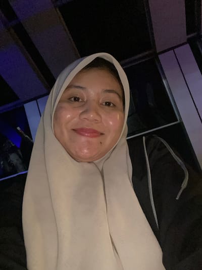 This is a close-up, self-portrait of a young woman wearing a light-colored hijab. Her face is turned towards the camera, and she is smiling. She appears to have a dewy complexion and is wearing light pink lipstick. She is wearing a dark-colored jacket, and the hijab is a pale beige color. The background is dark and out of focus, but it appears to be an indoor setting with some dark, geometric shapes and blue lighting, possibly indicative of a modern interior or a dimly lit public space. The image is oriented sideways. The time of day and weather are not discernible from the image. There is no visible text. The location context of Banjarbaru, Indonesia is provided.