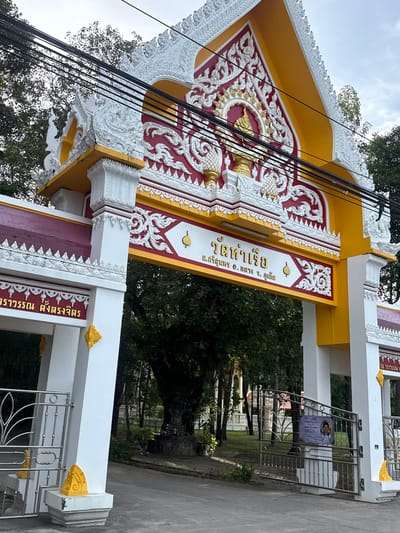 The image shows an ornate yellow and white arched gateway with intricate red and gold decorative carvings. Above the archway, a series of black electrical wires are strung across the frame. The gateway is likely an entrance to a temple or religious site, given its elaborate design and traditional motifs. Text in Thai script is visible on the gateway, identifying it as "วัดท่าเรือ" (Wat Tha Ruea). Below the main inscription, there is further Thai text indicating the temple's location as "ต.ศรีสุนทร อ.ถลาง จ.ภูเก็ต" (Tambon Si Sunthon, Amphoe Thalang, Phuket Province). This places the gateway in the Si Sunthon subdistrict of Thalang district, Phuket province, Thailand. The background consists of lush green trees and foliage, suggesting a tropical or subtropical environment. A metal gate with decorative vertical bars is partially visible to the left of the gateway, and a sign with an image of a person and text in Thai is attached to a similar gate on the right. The ground in front of the gateway appears to be paved with asphalt. The sky is overcast, with a pale, diffused light.