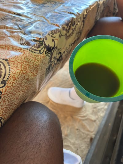 A green plastic cup filled with dark liquid sits above a patterned surface covered in clear plastic. Partially visible are two legs, one fair-skinned with a white sock and the other with dark skin. A metallic surface appears in the top right corner. The scene is captured from an overhead perspective.