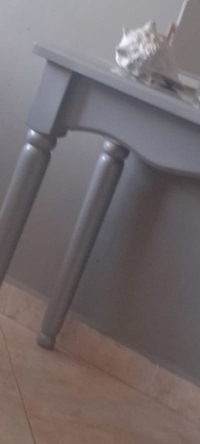 A close-up, low-angle, medium shot captures the legs and part of the tabletop of a gray console table. The table has a slightly curved apron. Two of its four legs are visible, both appearing cylindrical with rounded decorative elements midway down their length, leading to tapered feet that rest on a light brown tiled floor. Resting on the tabletop, to the right of the frame, is a white, amorphous object that appears to be a decorative shell or similar natural artifact. The background consists of a plain, muted gray wall, suggesting an indoor setting. The image does not provide any specific geographical indicators for Santa Ana, Venezuela.