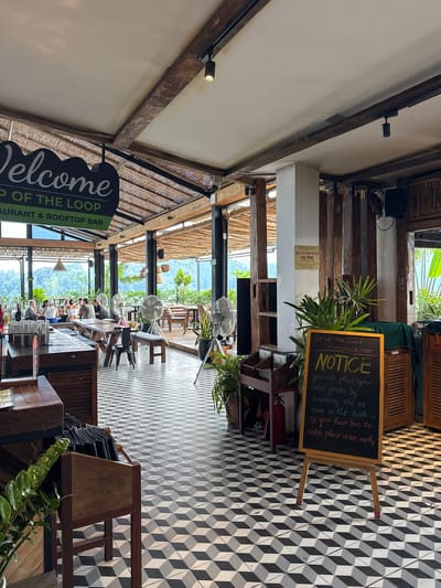 The image depicts the interior of "TOP OF THE LOOP RESTAURANT & ROOFTOP BAR" located in Hà Giang 1 Ward, Vietnam. The scene captures a spacious, open-concept dining area during the daytime, characterized by a rustic and natural design.
The setting features prominent wooden beams and columns supporting a textured, possibly woven bamboo or wicker, ceiling. Several white ceiling fans are suspended, suggesting a warm climate. The floor boasts a distinctive black, white, and grey geometric patterned tile that creates a three-dimensional cube effect. Lush green plants are interspersed throughout the venue, complementing the natural light streaming in from large windows or open sections along the far wall.
In the mid-ground and background, several people are seated at wooden tables and chairs, appearing to be dining or relaxing. Beyond them, a panoramic view of verdant mountains and dense tropical foliage is visible, emphasizing the scenic location.
Notable textual elements include a large green and white sign on the left that reads: "Welcome TOP OF THE LOOP RESTAURANT & ROOFTOP BAR." On the right, a black chalkboard easel displays a "NOTICE" in white chalk: "you can place your order faster by scanning the QR code on the table. If you have bus to catch please order early." Additionally, a smaller yellow sign mounted on a white wall section contains Vietnamese text: "CÔNG TY CỔ PHẦN ĐẦU TƯ DU LỊCH LÚA VÀNG CHI NHÁNH HÀ GIANG," which translates to "LUA VANG TOURISM INVESTMENT JOINT STOCK COMPANY HA GIANG BRANCH." The overall atmosphere is bright, open, and inviting, suggesting a popular spot for enjoying food and views.