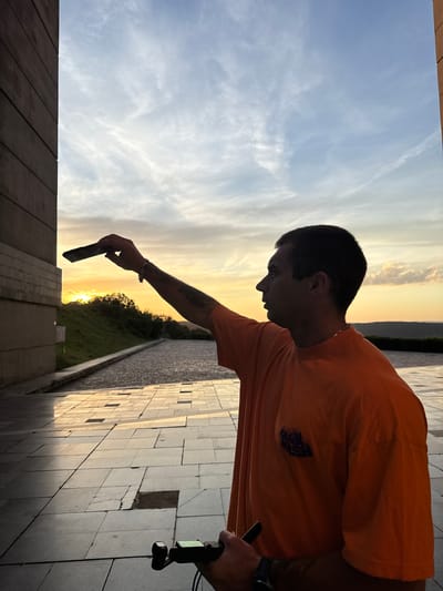 A young man wearing an orange t-shirt is standing on a paved outdoor area, holding a smartphone in his outstretched right hand, appearing to take a selfie. His body is partially silhouetted against a sunset sky with scattered clouds. To his left is a large, dark-colored building with a stone facade. The ground is paved with large, square tiles, some of which are cracked or missing. In the background, there is a grassy slope and distant hills. The scene is illuminated by the warm light of the setting sun. The image appears to have been taken at the Monument to the Founders of the Bulgarian State in Shumen, Bulgaria, due to the architectural style of the building and the monument's location overlooking the city.