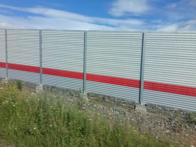 The image depicts a long, horizontal sound barrier constructed of corrugated metal sheeting, divided into several panels by vertical gray metal supports. A prominent horizontal red stripe runs across the lower portion of the barrier. The ground in front of the barrier is covered with gravel and loose stones. To the left and extending into the foreground is a dense patch of tall green grass with scattered wildflowers. The barrier is set against a backdrop of a blue sky with scattered white clouds. The overall scene appears to be an outdoor environment, possibly along a roadway or railway line, in or near Новый Мир, Russia. There are no people or active subjects visible in the image.