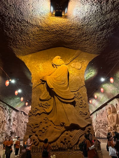The image displays a dramatically lit interior of a cave or grotto, featuring large, intricate stone carvings. The central subject is a massive horizontal relief sculpture, likely depicting a reclining Buddha or Bodhisattva, carved directly into the natural rock face. This monumental figure is rendered with flowing drapery and is surrounded by patterns resembling waves or clouds, all illuminated by a warm, golden spotlight that casts deep shadows, enhancing the three-dimensional quality of the carving.

The cave walls themselves are rough and textured, showing the natural rock formations. Above the main carving, a series of red, cylindrical lanterns are strung across the ceiling, providing a soft, ambient red glow, while other spotlights highlight different sections of the cavern, some casting a greenish tint on parts of the rock.

On the right side of the image, another section of the cave wall features a multitude of smaller, detailed relief carvings depicting various figures, suggesting a narrative or a collection of deities. Several visitors, dressed in modern attire, are visible on a platform in the lower right, observing the ancient artwork. Some individuals are looking up at the carvings, and one person stands with their hands clasped, indicating a moment of contemplation or reverence. A white tote bag carried by a visitor features East Asian characters. Near the group, a sign is partially visible with Chinese characters that read "注意安全" (Pay attention to safety) and possibly "小心台阶" (Mind the steps).

The artificial lighting creates a mystical and atmospheric setting, emphasizing the age and spiritual significance of the grotto. The scene captures an experience of cultural exploration within a historic and sacred space.