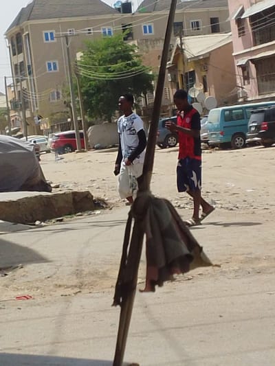 The image depicts a daytime street scene in Sabon Gari East, Nigeria. In the immediate foreground, a dark vertical pole with a collapsed, dark, tattered fabric object (possibly an umbrella) attached to it occupies the lower center. Two males are walking from left to right across the midground. The individual on the left wears a white t-shirt with dark patterns, black long sleeves, and light-colored shorts, looking straight ahead. The individual on the right wears a red and black short-sleeved shirt with a white logo, blue and white patterned shorts, and white slide sandals; he is observed looking down at a mobile phone held in both hands. The ground consists of uneven dirt and paved sections. In the background, a light-colored multi-story building with blue-framed windows and a water tank on its roof is visible on the left, while other buildings, including one with satellite dishes, line the right. Several vehicles, including a red SUV, a light blue van, and a dark SUV, are visible parked or in motion further down the street. The scene is brightly lit by direct sunlight.