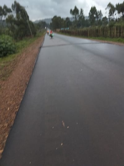 The image depicts a rural or semi-rural road scene in Nyansiongo ward, Kenya, under an overcast sky, suggesting low light conditions such as dusk, dawn, or a heavily clouded day. The paved road surface appears dark and possibly damp or wet. To the left, a reddish-brown dirt shoulder abuts the asphalt, followed by dense green vegetation including bushes and trees. In the distance, two individuals are visible on the road, moving away from the viewer. The lead person is on a bicycle, displaying a visible red or orange item. Behind them, a second individual is also on a bicycle, carrying a large, light-colored, possibly green or light blue, bundle or object. Further down the road on the left, a distinct white light source is apparent. The right side of the road in the mid-ground features a long, low wooden fence or barrier, behind which more trees are visible. Distant hills or mountains form the horizon line on the far left.