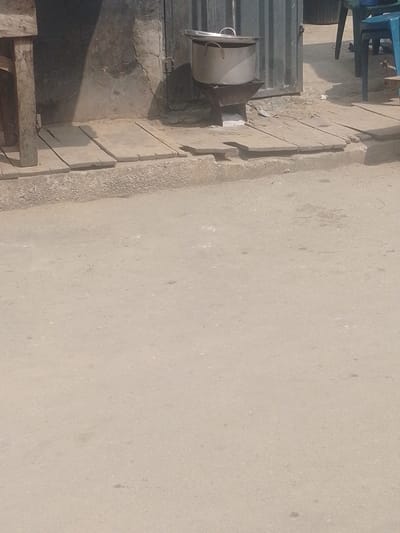 The scene is a ground-level view of an outdoor urban environment in Agani, Nigeria, during daylight. The foreground consists of a light-colored, possibly unpaved dirt or gravel road surface with minor irregularities and debris. In the midground, a raised pedestrian path constructed from multiple light-colored wooden planks runs horizontally across the frame, forming a transition from the road to a building structure.
On this wooden plank path, a large, silver-colored aluminum pot with a matching lid rests on a dark-colored, rudimentary metal cooking stand or charcoal stove. This stove is positioned directly in front of a weathered, light-colored wall exhibiting visible cracks and damage. Immediately to the right of the wall section is a dark, corrugated metal door or gate, which appears slightly ajar. To the far left, a vertical wooden beam, likely part of a table or stall structure, is partially visible. Further to the right in the background, a portion of a blue plastic chair and a dark, indistinct object are present. No individuals are depicted in the frame.