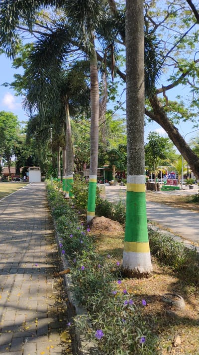 The image depicts a sunlit park scene in Madiun, Indonesia, likely a public recreational area. A paved walkway, constructed of grey interlocking bricks, runs diagonally from the lower left to the middle ground, bordered by a low concrete curb. Along the right side of the walkway, a row of palm trees stands, their trunks painted with alternating bands of green and yellow near the base. Purple flowers bloom in lush greenery adjacent to the walkway and around the base of the trees.

In the background, beyond the trees, the park continues with more greenery and a few low-lying structures, possibly kiosks or small buildings. A sign is visible in the distance, reading "MINI PICELAND". People and vehicles are present in the far background, suggesting a lively atmosphere. The sky is a clear, bright blue, indicating a sunny day. The overall impression is of a well-maintained park on a pleasant day.
