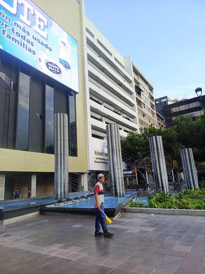 The image depicts a street scene in Guayaquil, Ecuador, likely in an urban commercial area. In the foreground, a man wearing a white, red, and blue striped shirt, blue jeans, and a white baseball cap walks on a paved area, holding a yellow and blue toy horn. He is walking towards the right side of the frame.

In the mid-ground, there is a water feature with several tall, fluted pillars through which water sprays. Behind the water feature, a building with large windows displays a prominent advertisement for "ZOTE" soap and detergent on a large digital screen. The advertisement features images of Zote products and text in Spanish, including "rón más usado por todas familias" (most used by all families). Adjacent to the Zote advertisement, another building is visible, with multiple floors and windows. Further back, there are signs for McDonald's.

In the background, several other buildings of varying heights and architectural styles are visible against a clear blue sky. Some of the buildings appear to be modern office buildings or residential complexes. The overall atmosphere suggests a daytime scene in a bustling city.