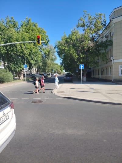 The image depicts a street scene in Astana, Kazakhstan, under a clear blue sky. In the foreground, the rear of a white car is visible on the right. The street features a pedestrian crossing. Three individuals are crossing the street from left to right: two younger people, one wearing a white t-shirt and a backpack, and another in a dark t-shirt and shorts, both with hats; and a third person, further ahead and to the right, wearing a light blue outfit and carrying a white bag. A traffic light is visible on a pole, showing a red light. Traffic signs are also present, including a blue sign with a white "P" indicating parking, and a blue sign with a white upward-pointing arrow. Trees line the street, and a modern building with a light-colored facade is visible on the right side of the frame. The overall environment suggests a sunny day in an urban setting.