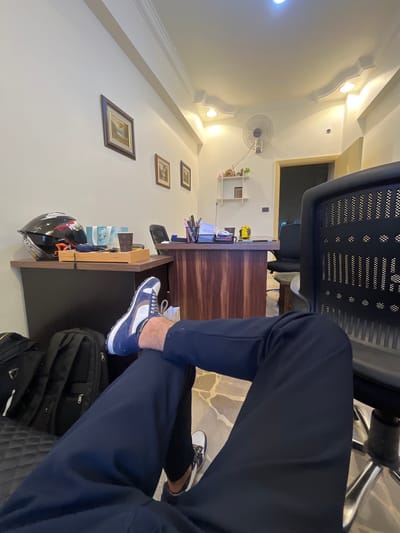 The image depicts an indoor scene, likely an office or study, in Rukn ad-Din Municipality, Syria. The perspective is from a seated individual whose legs and feet are prominently featured in the foreground, clad in dark blue trousers and white and blue athletic shoes. The individual's left leg is crossed over the right.
The environment includes a dark-colored office chair with a perforated backrest in the right foreground. Behind the chair, a wooden desk with a dark brown, textured front is visible, laden with office supplies such as pens in a holder, notebooks, and a yellow object. A white shelf unit is mounted on the wall to the left of the desk, displaying a few small decorative items.
Further to the left, a dark desk surface holds a black helmet with red and white accents, along with a small wooden tray containing several blue-wrapped items and a brown disposable cup. Two black backpacks are placed on the floor to the left of this desk.
The walls are painted a light color, and decorative molding is present along the ceiling. A ceiling-mounted fan is visible on the wall. Several framed pictures, appearing to be landscape paintings, are hung on the wall. The floor is tiled with a pattern of light and darker stones. The overall lighting suggests an indoor environment with artificial light sources.