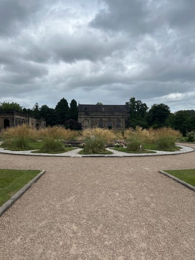 The image shows a view of a historic building, likely a church or cathedral, with intricate stone architecture. The building is surrounded by lush green grass, mature trees, and decorative ornamental grasses. A pathway made of gravel is prominent in the foreground, curving to the right and bordered by grass and stone. The sky is overcast with dramatic, heavy grey clouds, suggesting a cool or temperate climate.
The scene appears to be a well-maintained garden or park area adjacent to the historic structure. There are no people visible in the image, and the activity taking place is simply the presentation of the landscape and architecture. Notable details include the textured stone of the building, the feathery appearance of the ornamental grasses, and the light-colored gravel path. The overall mood is serene and somewhat somber due to the cloudy sky.
The location context is Stafford, United Kingdom, which aligns with the architectural style and the greenery. There is no visible text in the image. The time of day is difficult to ascertain precisely due to the overcast conditions, but it appears to be daytime.