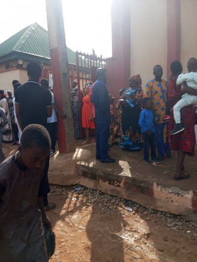 This image depicts an outdoor gathering of people in what appears to be the city of Narayi, Nigeria. The scene is characterized by a mix of individuals, including adults and children, some of whom are dressed in vibrant, patterned clothing, suggesting a cultural or festive event. In the foreground, a young boy wearing a dark garment is looking downwards, holding a dark bag. Behind him, other people are standing and conversing, with some children visible in the mid-ground, including a child in a blue shirt being held by an adult. Further back, a group of adults are gathered, some in traditional attire. The background shows a building with a green roof, a fence, and a wall. The ground is a mix of dirt and gravel, with some debris scattered around. The lighting suggests it is daytime, with the sun casting shadows. The overall impression is of a community event or gathering in an urban Nigerian setting.