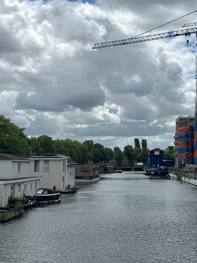 The image depicts a canal scene in Amsterdam, Netherlands, with a cloudy sky. In the foreground, the water of the canal is calm. On the left bank, several white houseboats are moored, one with a black boat docked next to it. Further down the canal, on the right bank, a blue structure with "DWM" branding is visible, adjacent to a multi-story building under construction, with scaffolding and orange and blue sheeting. Trees line the background on the left bank, and a bridge crosses the canal in the distance. A construction crane is visible in the upper right quadrant of the image, against the cloudy sky.