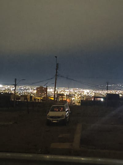 The image depicts a nighttime scene in the city of Arica, Chile, looking out over a densely populated area illuminated by numerous lights. In the foreground, a white van is parked on a dirt road. The van's interior lights are on, and it appears to be occupied by at least two people. The background shows a sprawling urban landscape with buildings and lights stretching across the horizon. The sky is dark and cloudy. Various utility poles and wires are visible in the midground, silhouetted against the distant city lights. The overall impression is one of a residential area at night, possibly on a hillside overlooking the city.