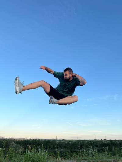 A man is captured mid-air, performing a martial arts move against a clear blue sky. He wears a green t-shirt and black shorts, with grey athletic shoes on his feet. His body is positioned horizontally, with his right leg extended forward and his left leg bent. His right arm is outstretched in a punching motion, while his left arm is bent and held near his head. Tattoos are visible on his arms. The background consists of a vast expanse of blue sky, with a faint horizon line of trees and distant structures visible at the bottom of the frame. The location is Shumen, Bulgaria.